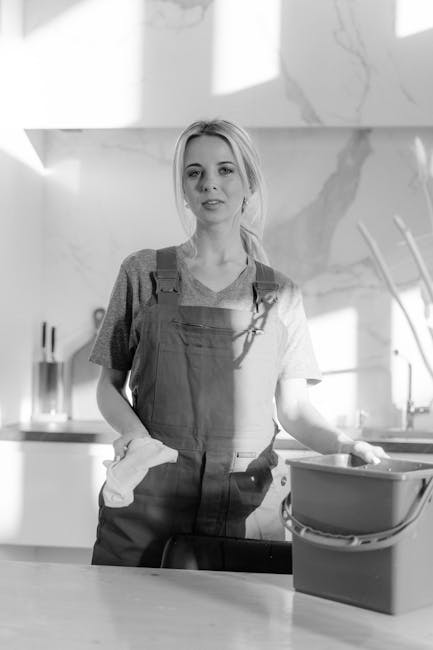 A woman wearing a grey t-shirt and red apron is engaged in surface cleaning within a modern kitchen. She stands beside a white countertop, which appears to be made of a smooth material such as quartz