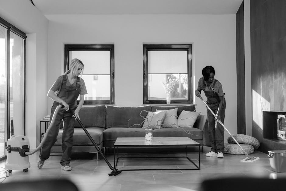 Two professional cleaners from Kennington Cleaner wearing orange overalls and grey shirts are performing surface cleaning in a modern living room. One cleaner is using a yellow vacuum cleaner to clean