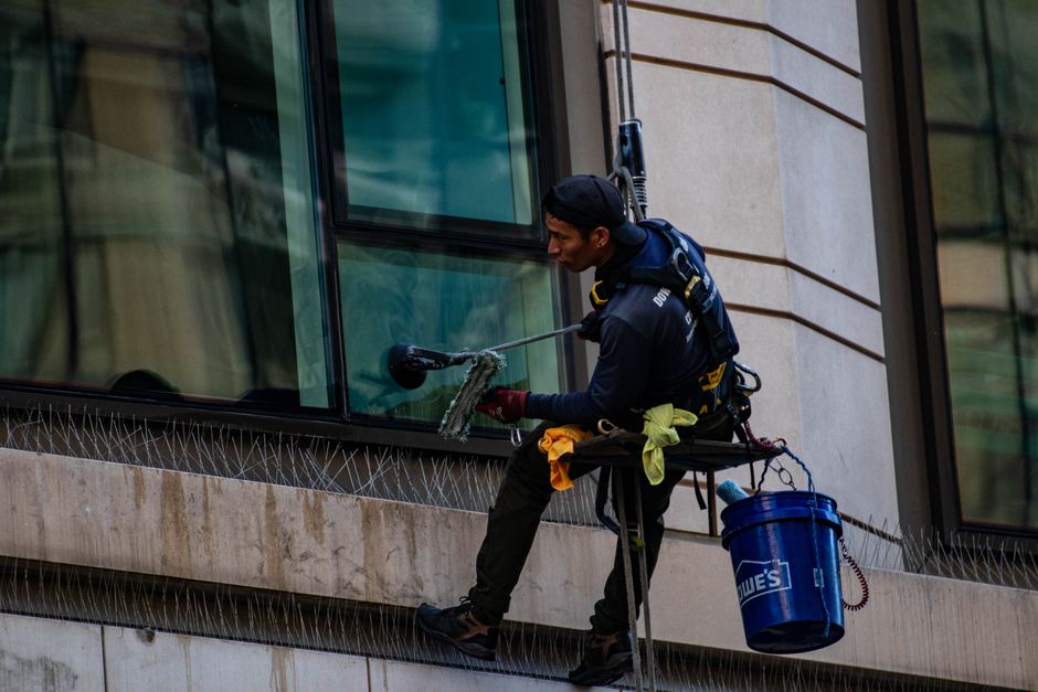 Two professional window cleaners from Kennington Cleaner are suspended on a secure harness system, working on the exterior of a high-rise building with large glass windows and a brick facade. They are