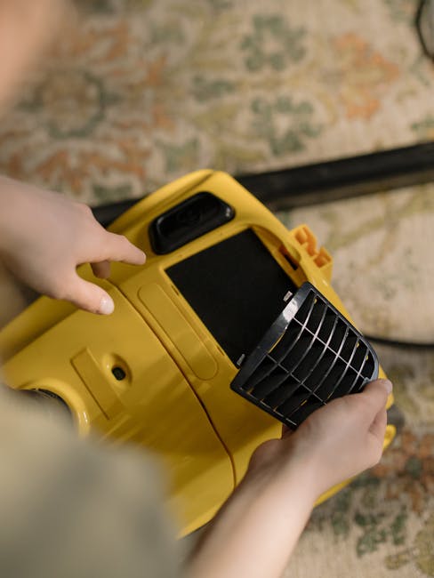 A man with gray hair and a beard is engaged in floor cleaning at a residential living room, using a canister vacuum cleaner with a hose attachment. The room features a hardwood parquet floor partially