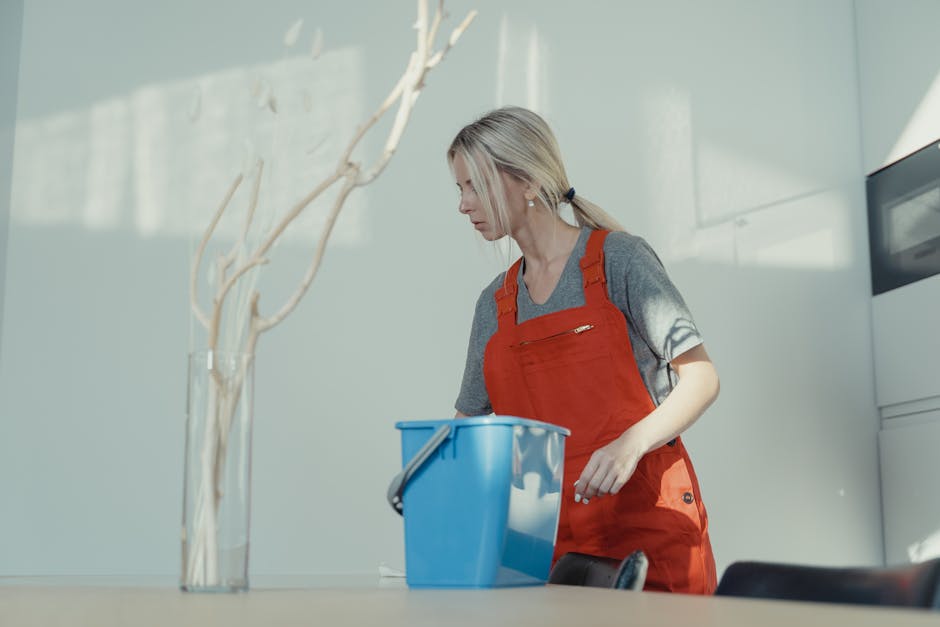 A woman with blonde hair tied back, wearing a grey t-shirt and a bright red cleaning apron, is engaged in surface cleaning inside a modern kitchen. She is standing and looking down at a blue cleaning bucket placed on a white countertop. The kitchen features a glossy white tiled backsplash, sleek cabinets, and a stainless steel oven on the right side. Sunlight streams through a nearby window, illuminating the workspace and highlighting the clean, dust-free surfaces. The scene captures a moment of domestic cleaning, emphasizing hygiene and meticulous attention to detail, consistent with the Kennington Park house cleaning guide for landlords, provided by Kennington Cleaner.