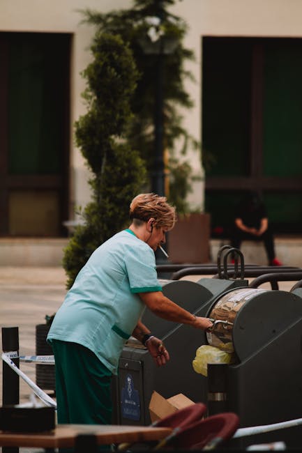 A person wearing a wide-brimmed hat, gloves, and a light blue jacket is engaged in surface cleaning or sorting waste on a paved street, surrounded by large plastic bags filled with waste materials. Th